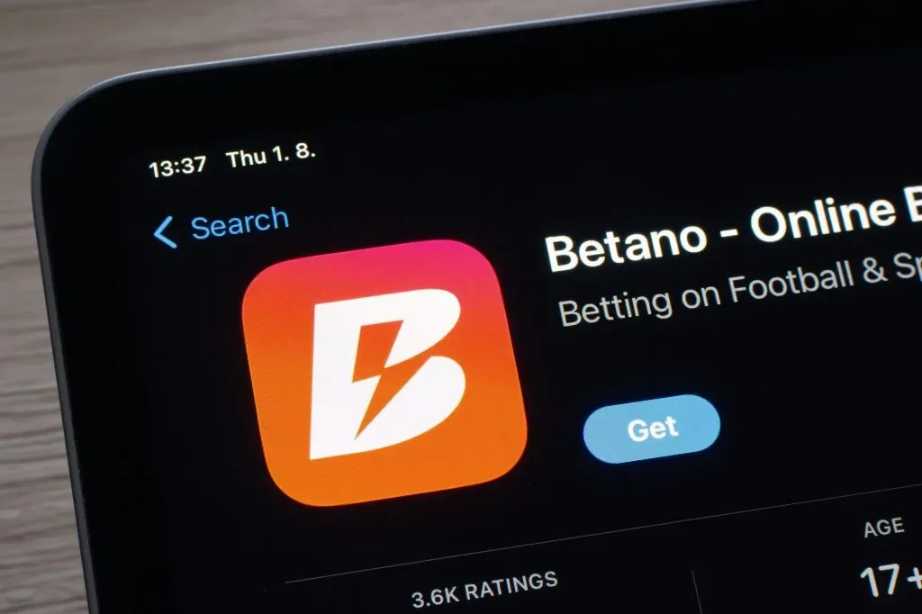 Primer plano en diagonal de una pantalla con la ficha de la aplicación de la marca en una tienda de apps: ícono naranja con un rayo, botón de descarga y, al pie, el recuento de valoraciones y la clasificación por edad; fotografía de un portal de guías.